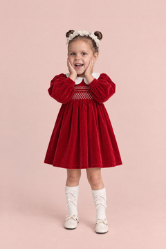 Holly Red Velvet Christmas Dress, front: a girl with both hands on her cheeks in the red velvet dress with a white Peter Pan collar and smocked bodice