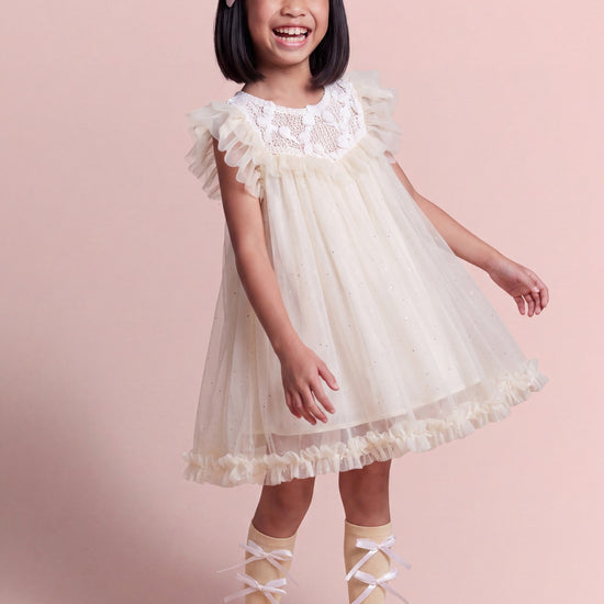 Girl wearing the Primrose ivory flower girl dress, white crochet yoke scattered with tiny gauze roses over sheer glitter tulle
