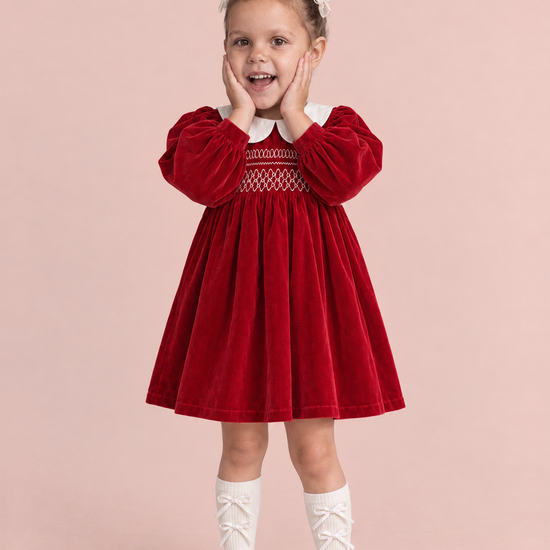 Holly Red Velvet Christmas Dress, front: a girl with both hands on her cheeks in the red velvet dress with a white Peter Pan collar and smocked bodice