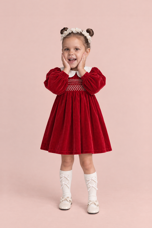 Holly Red Velvet Christmas Dress, front: a girl with both hands on her cheeks in the red velvet dress with a white Peter Pan collar and smocked bodice