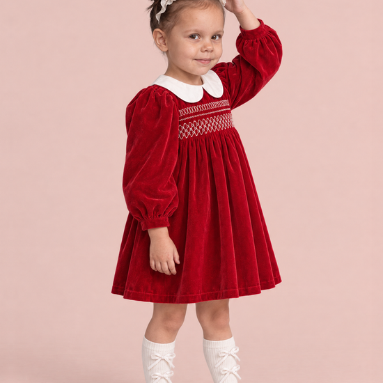 Holly Red Velvet Christmas Dress, three quarter view, one hand on her headband, showing the puff sleeves and the embroidered smocking