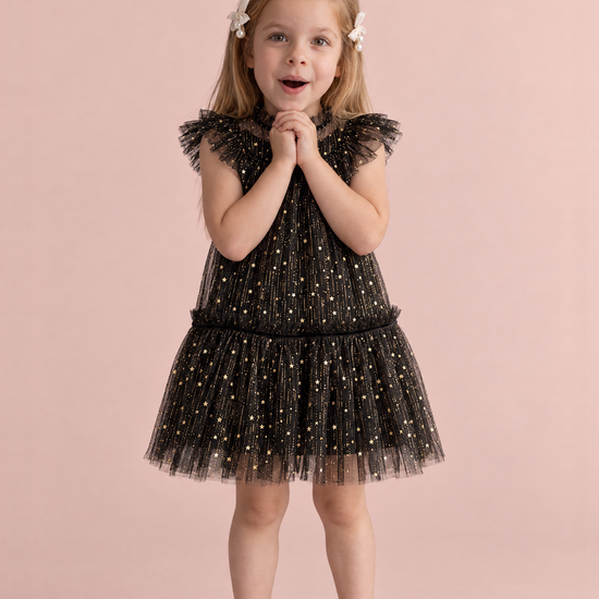 Stardust Black Star Tulle Party Dress, front: a girl with her hands clasped under her chin in the black tulle dress with gold stars and flutter sleeves