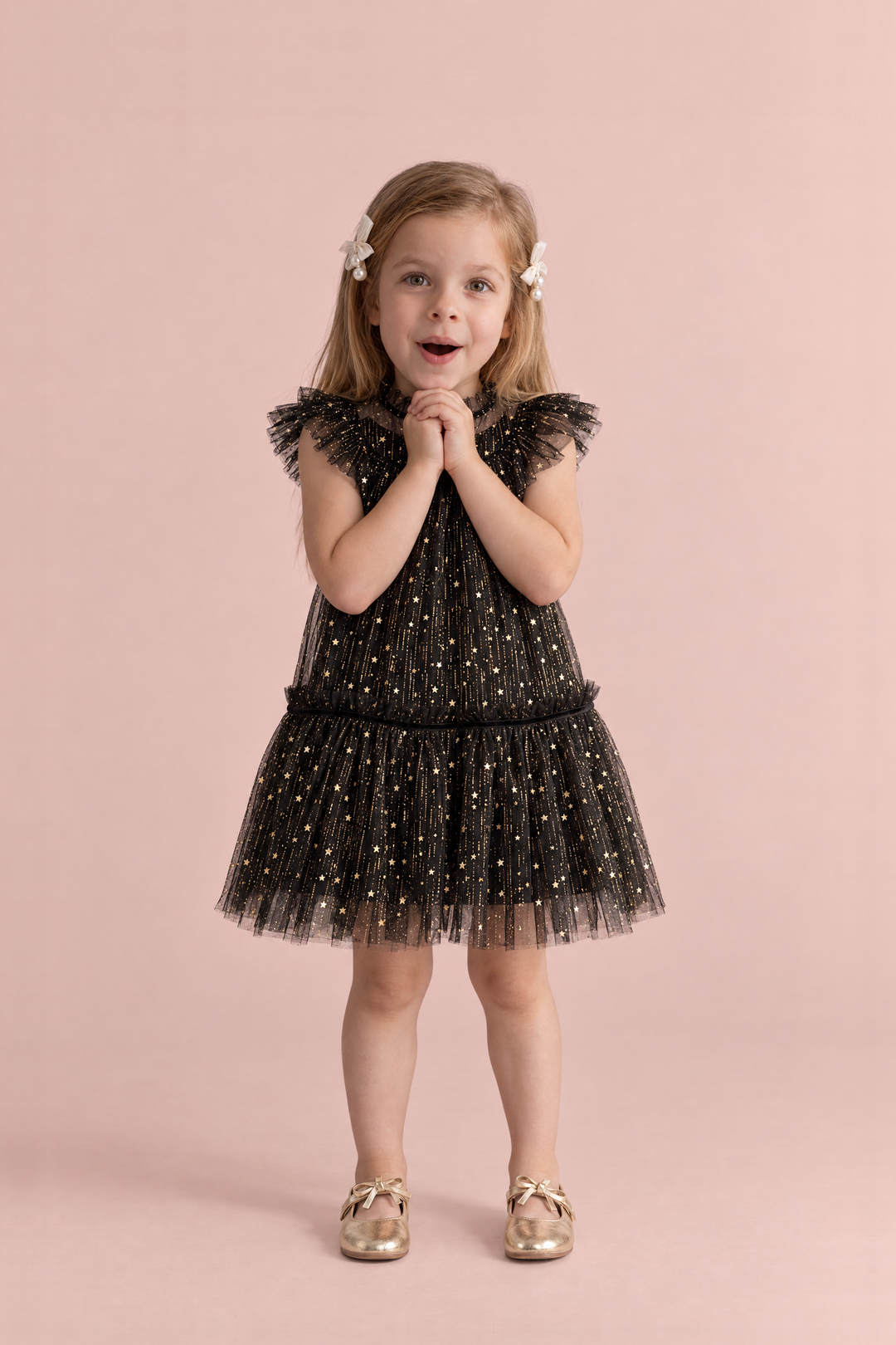 Stardust Black Star Tulle Party Dress, front: a girl with her hands clasped under her chin in the black tulle dress with gold stars and flutter sleeves