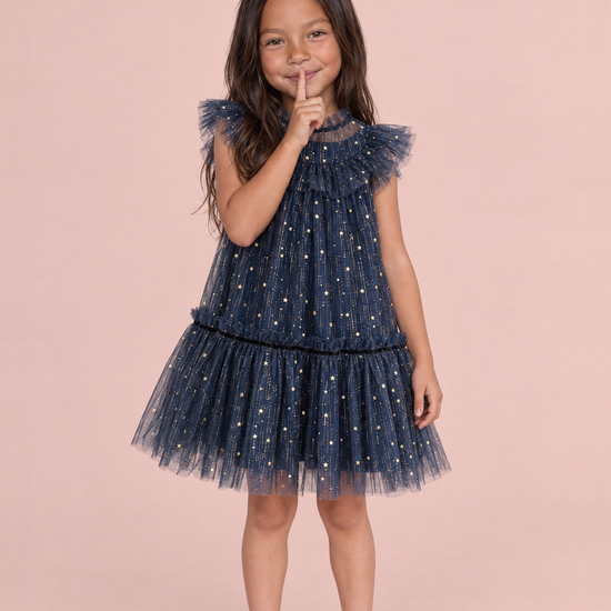 Stardust Navy Star Tulle Party Dress, front: a girl with a finger to her lips in the navy tulle dress with gold stars and flutter sleeves