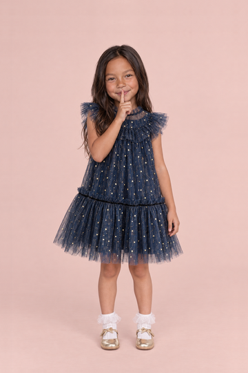 Stardust Navy Star Tulle Party Dress, front: a girl with a finger to her lips in the navy tulle dress with gold stars and flutter sleeves